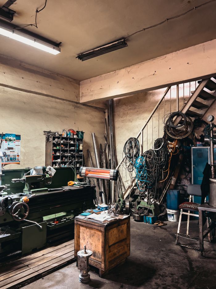 A cluttered industrial workshop featuring tools, machinery, and storage with cables and stairs.